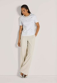 Witte shirt met korte mouwen en een bewerkte kraag, gecombineerd met beige wijde broeken en zilveren hoge hakken, tegen een effen achtergrond.