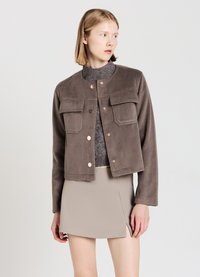 Veste en polaire marron avec un col rond, deux poches avant et fermeture à pression. Portée par-dessus un haut en maille gris et une jupe beige.