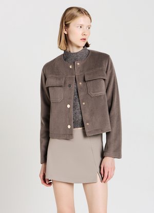 Veste en polaire marron avec un col rond, deux poches avant et fermeture à pression. Portée par-dessus un haut en maille gris et une jupe beige.