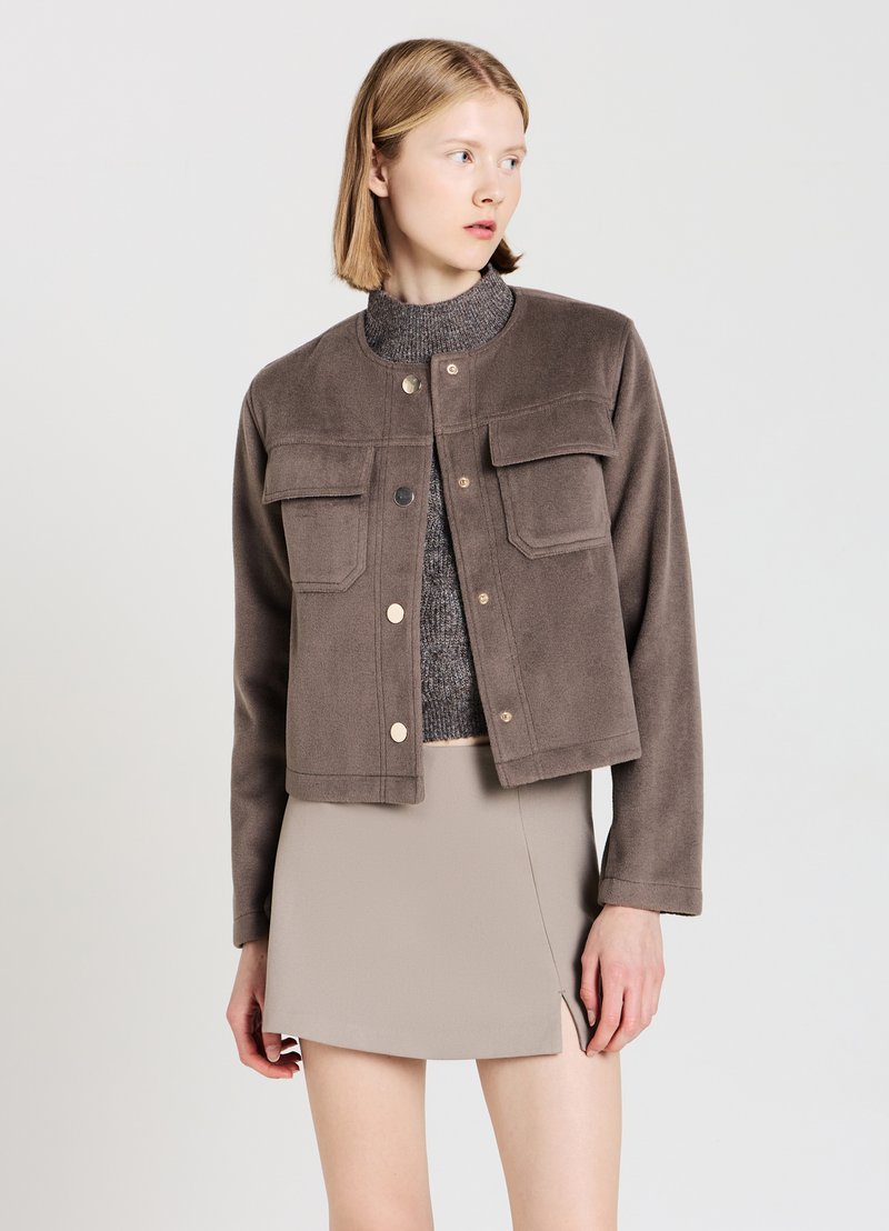 Veste en polaire marron avec un col rond, deux poches avant et fermeture à pression. Portée par-dessus un haut en maille gris et une jupe beige.