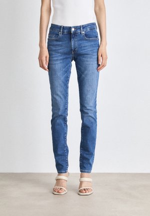 Slim fit blå denim jeans båret af en person, der står, kombineret med hvide åbne højhælede sandaler og en hvid ærmeløs top.