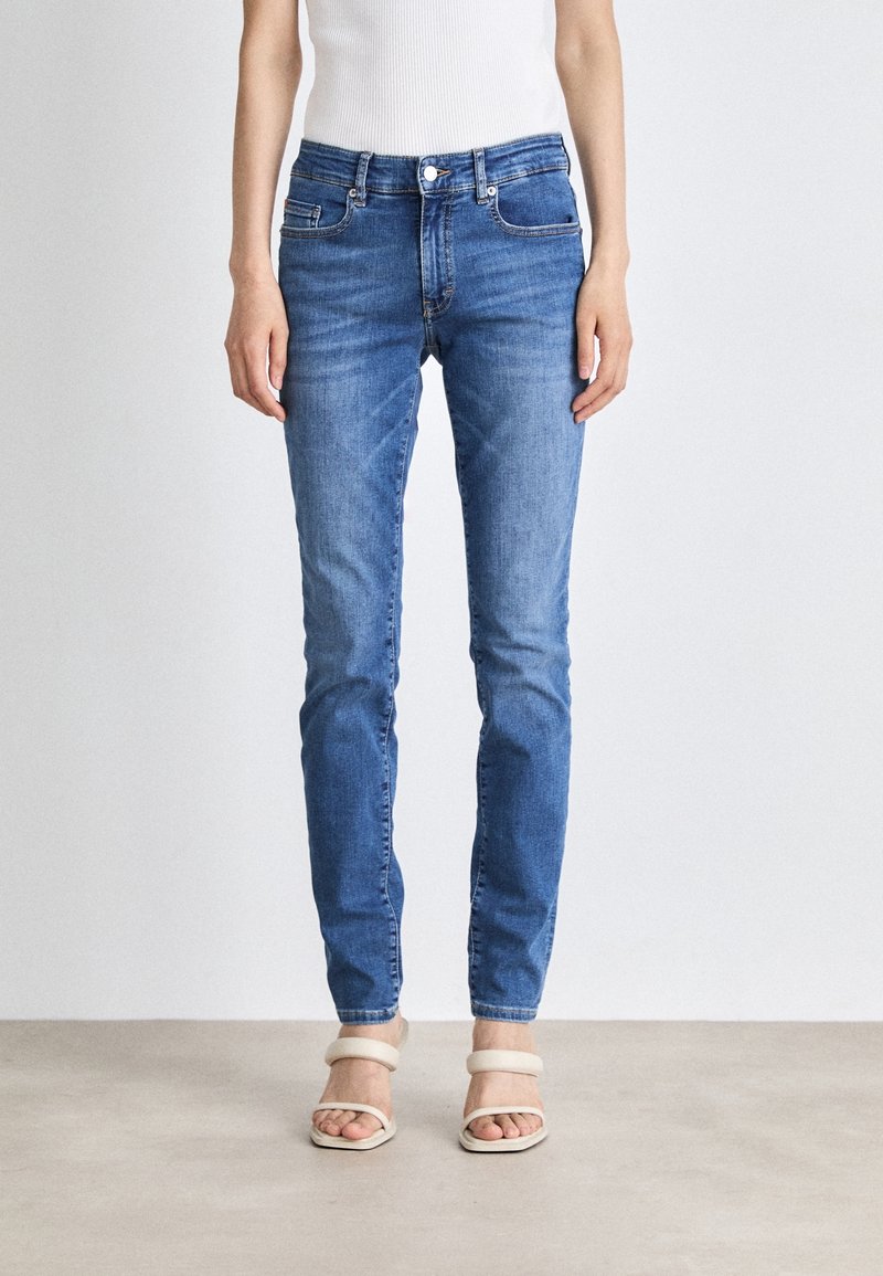 Jean slim fit bleu en denim porté par une personne debout, associé à des sandales à talons blanches à bout ouvert et un top blanc sans manches.