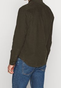 Mörkgrön manchesterkjorta med knäppkrage, söm vid bakre axelpartiet och rundad fåll. Kombinerad med blå jeans.