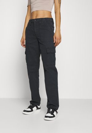 Personne portant un pantalon cargo noir, des baskets noir et blanc et un top court beige, debout devant un fond clair uni.