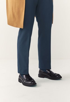 Unterschenkel mit marineblauer Hose, schwarzen Lederschuhen und einem camelbraunen Mantel, der seitlich vor einem einfarbigen Hintergrund drapiert ist.