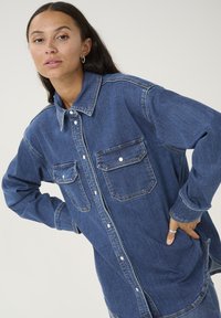 Tumesinine denim-särk, nööpidega, kahe rindkere taskuga, veidi laiem sobivus, tekstuuriga kangas, kontrastsed õmblused ja klassikaline krae kujundus.