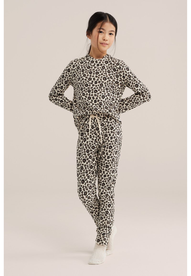 Fille debout, les mains sur les hanches, portant un haut et un pantalon assortis à manches longues avec un imprimé léopard noir et beige et des chaussettes blanches.