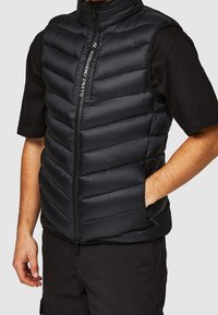 Sort puffervest med lynlås og høj krave. Har vandrette syninger, to sidelommer og en glat, glinsende overflade.