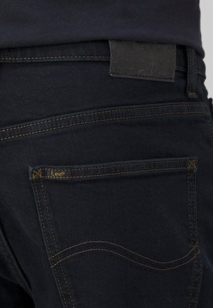 Primo piano della tasca posteriore di un jeans blu scuro con cuciture gialle e piccola etichetta del marchio Lee, indossato da una persona con camicia scura.