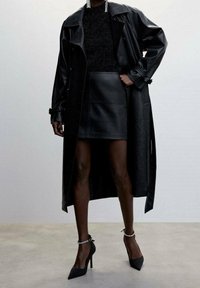 Černý kožený trench coat s převislým límcem, padnoucí černý svetr a černá sukně s vysokým pasem. Černé vysoké podpatky s kotníkovými řemínky.