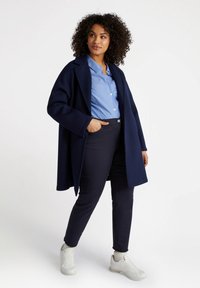 Tummansininen takki, pitkät hihat, avoin etuosa. Vaaleansininen raidallinen paita alla, yhdistettynä tummiin housuihin ja valkoisiin lenkkareihin. Sujuva pinta.