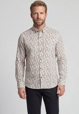 Blonde man die een lang mouwig, knoopjes overhemd draagt met een subtiel meerkleurig abstract patroon en een zwarte broek, staande voor een grijze achtergrond.