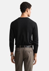 Pull en maille noire à col rond, manches ajustées, associé à un pantalon marron à motifs. Le modèle est debout, de dos par rapport au spectateur.