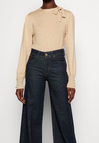 Beige långärmad topp med rosett på axeln och knäppta manschetter, kombinerad med högmidjade vida mörk denimjeans på en stående person.