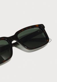 Occhiali da sole tartaruga marrone con lenti verde scuro. Design della montatura rettangolare con finitura lucida e branding minimale sulle lenti.
