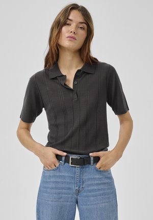 Kobieta w ciemnej, krótkiej koszulce polo wpuszczonej w jasnoniebieskie dżinsy z czarnym paskiem, stojąca z rękami w kieszeniach.