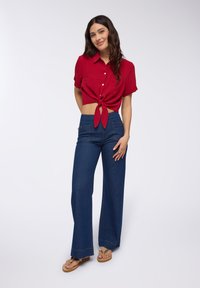 Camicia rossa corta con bottoni e fiocco frontale, abbinata a jeans a gamba larga blu scuro a vita alta. Modella che indossa sandali piatti color beige.