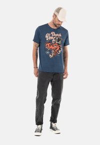 T-shirt en coton bleu avec un graphique orange audacieux d'un dragon et le texte "Dutch Von" sur le devant, associé à un jean noir et des baskets.