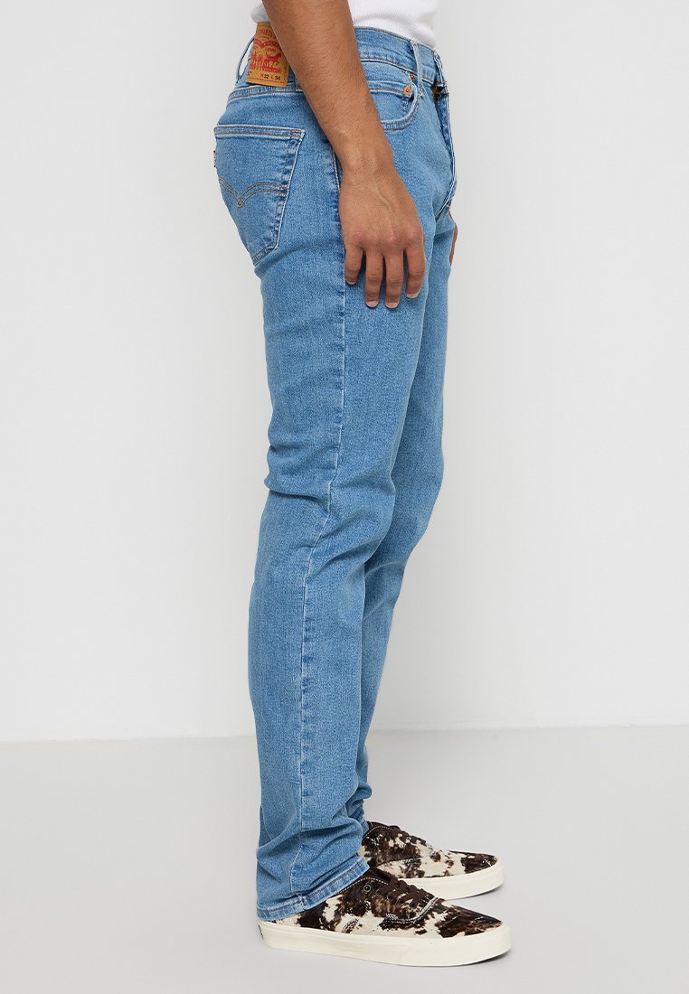 Jean en denim bleu clair avec une coupe slim, arborant une étiquette en cuir marron à l'arrière et un léger délavage. Associés à des sneakers à motifs.