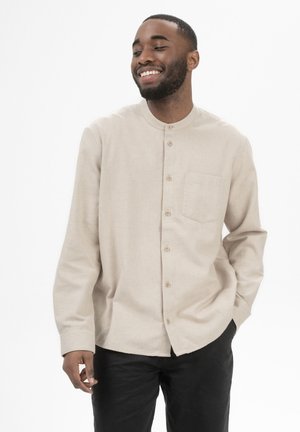 Lachende man met korte baard, gekleed in een beige overhemd met knopen en zwarte broek, staand met één hand in de zak tegen een witte achtergrond.