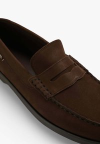 Mocasines de ante marrón con un detalle de costura, una barra elevada en la parte superior y una suela de goma plana. Textura suave con acentos mínimos.