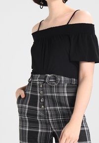 Černý top bez ramínek s nařaseným okrajem v kombinaci s černobílými kostkovanými kalhotami s vysokým pasem, které mají pásek a detail knoflíku.