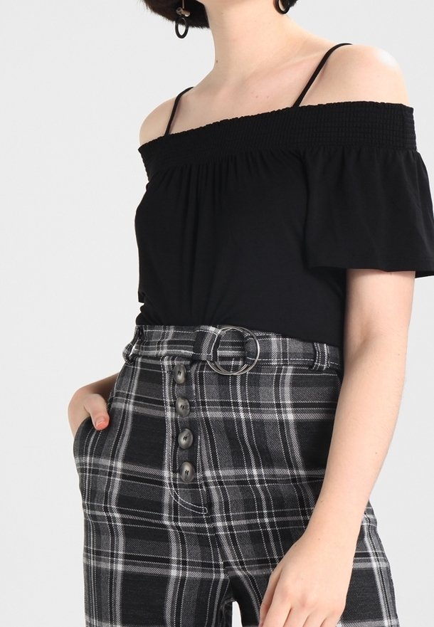 Černý top bez ramínek s nařaseným okrajem v kombinaci s černobílými kostkovanými kalhotami s vysokým pasem, které mají pásek a detail knoflíku.