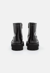 Botas de tobillo de charol negro con dos cremalleras laterales, suelas de goma gruesa y detalles de costura blanca.