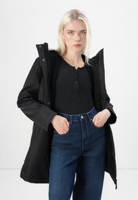 Black waterproof coat with fur lining, featuring a hood and snap buttons. Paired with a black ribbed top and high-waisted denim jeans.