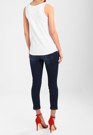 Weißes ärmelloses Oberteil mit taillierter Passform, kombiniert mit dunkelblauen Skinny Jeans und roten High Heels, die Knöchelriemen und offene Rückseiten zeigen.