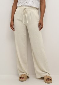 Pantaloni largi de culoare bej deschis, confecționați dintr-un material texturat, având un elastic în talie și șnur pentru ajustare. Asociați cu sandale slip-on împletite de culoare maronie.