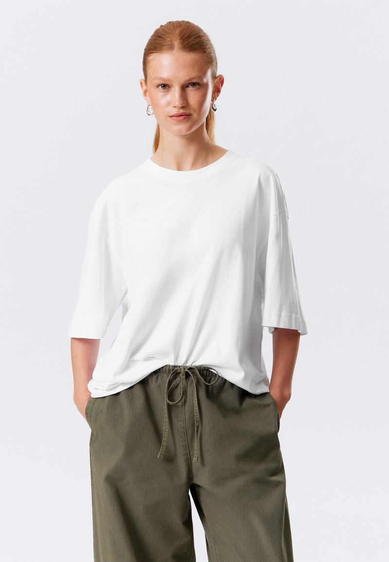 Weißes Baumwoll-T-Shirt mit kurzen Ärmeln und Rundhalsausschnitt; kombiniert mit lockeren olivgrünen Hosen mit Kordelzug und seitlichen Taschen.