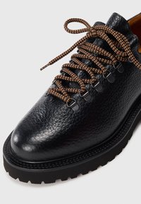 Black leather shoe with a textured finish, featuring brown and black patterned laces, metal eyelets, and a rugged black outsole.