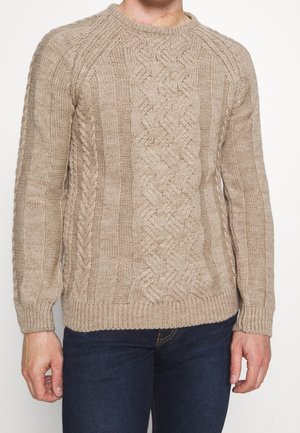 Homme portant un pull en laine beige à torsades et un jean bleu foncé, debout devant un fond blanc uni.