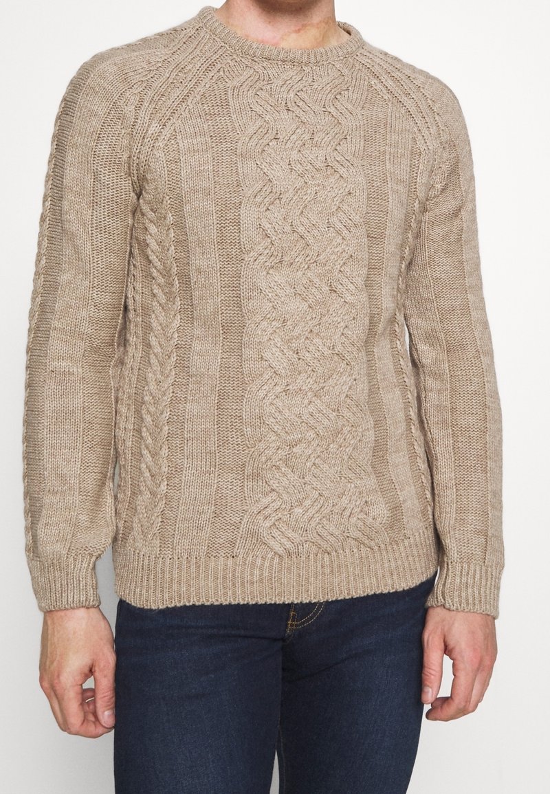 Homme portant un pull en laine beige à torsades et un jean bleu foncé, debout devant un fond blanc uni.
