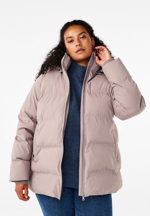 Eine Pufferjacke in hellem Beige mit hohem Kragen, Reißverschluss und gestepptem Muster. Tragen über einem navyfarbenen Pullover und blauen Jeans.
