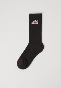 Calza nera lavorata a maglia con texture a costine, presenta un ricamo di design di sneaker bianche e testo rosso vicino alla punta. Lunghezza standard alla caviglia.