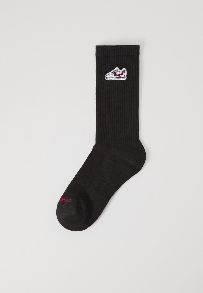 Calza nera lavorata a maglia con texture a costine, presenta un ricamo di design di sneaker bianche e testo rosso vicino alla punta. Lunghezza standard alla caviglia.