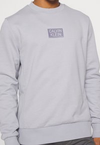 Ljuslila sweatshirt med ribbade muddar och fåll, med en rektangulär logotypmärke på bröstet i mörkare lila. Mjuk tyg, rund halsringning.