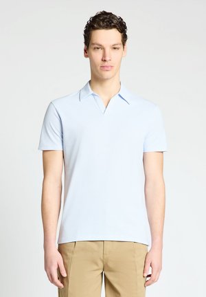 Jeune homme aux cheveux courts et bouclés portant une chemise bleu clair à manches courtes avec col et un pantalon beige, debout devant un fond uni.