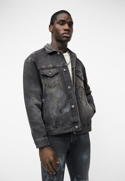 Άνδρας που φοράει σκουρόχρωμο γκρι τζιν μπουφάν με μοτίβο και τζιν παντελόνι που ταιριάζει, στέκεται με το ένα χέρι στην τσέπη, μπροστά από απλό λευκό φόντο.