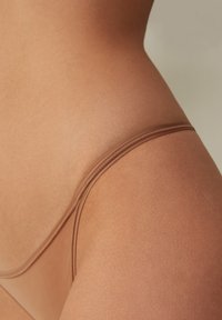 Beige stringslips i ett mjukt och elastiskt tyg. Har tunna elastiska band och en ren finish längs kanterna.
