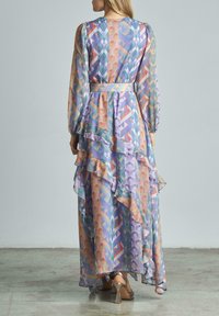 Langes, mehrfarbiges Kleid aus gemustertem Stoff, mit Rüschenschichten und langen Ärmeln. Besitzt einen Gürtel in der Taille; der transparente Stoff sorgt für Textur.