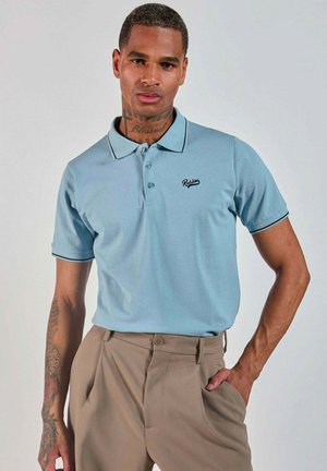 Homme tatoué portant une chemise polo bleu clair des Redskins et un pantalon beige, se tenant avec une main dans la poche sur un fond uni.