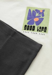 T-shirt blanc avec un graphique coloré de fleurs en bleu et orange, accompagné d'un texte noir disant "BONNE VIE LIBÈRE TON ÂME". Manches côtelées grises.