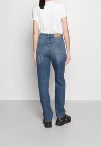 Person som står vänd bort, iförd en vit t-shirt och blå jeans, med svarta skor. Bakgrunden är klar och ljus.