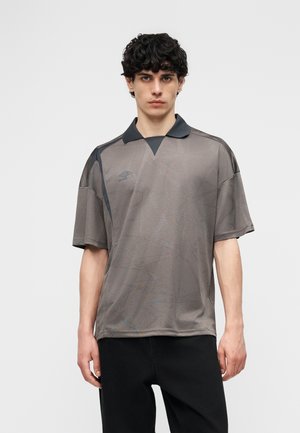 Młody mężczyzna ubrany w luźną, szarą sportową koszulkę z subtelnymi geometrycznymi wzorami i czarnym kołnierzem, stojący na tle jednolitego jasnego tła.