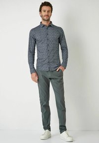 Langärmliges marineblaues Hemd mit einem weißen geometrischen Muster, kombiniert mit hellgrauen Hosen und weißen Sneakers. Das Hemd hat einen Button-Down-Kragen.