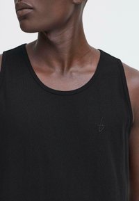 Svart ermeløs tanktopp laget av glatt stoff, med rund halslinning og subtilt brodert logo på brystet.