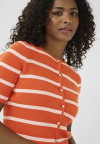 Cardigan tricoté orange à manches courtes avec des rayures blanches, présentant un col rond et des accents de boutons dorés sur le devant.